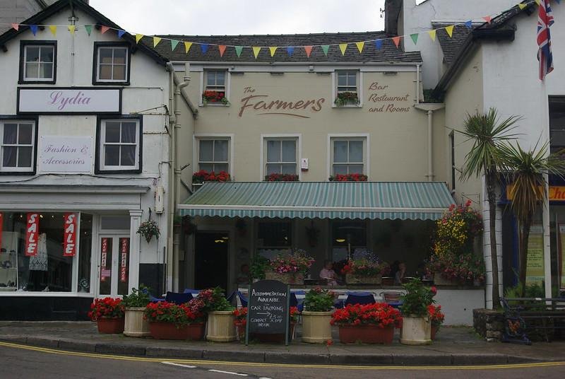 A pub and restaurant with a striped green and white canopy, floral displays, and a chalkboard sign advertising accommodations. Adjacent boutique with "SALE" signs and bunting overhead.