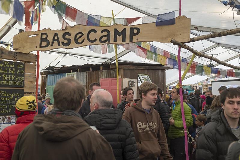 Basecamp, Kendal Mountain Festival | A crowded indoor festival setting with people gathered under a tent. A wooden sign reading "BASECAMP" is hanging overhead, and various colourful flags are strung across the space.