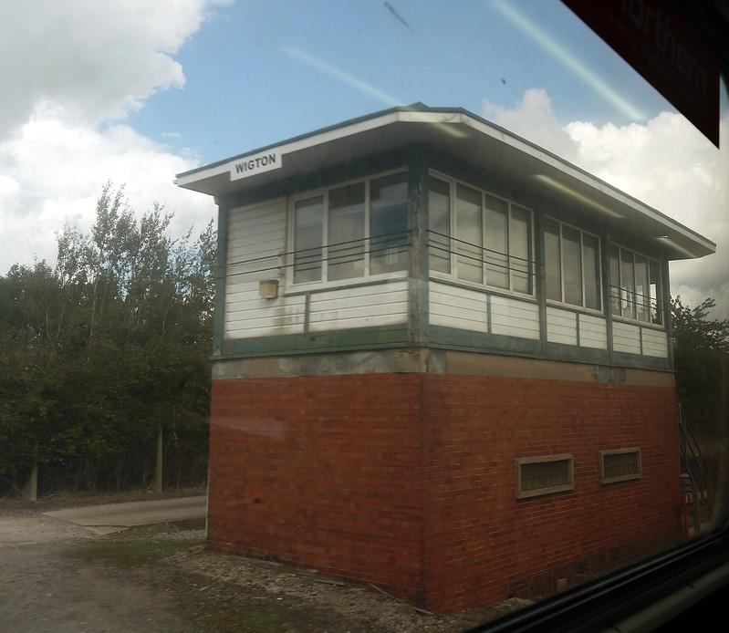 Wigton signal box | A red brick signal box with "Wigton" sign, viewed from a moving vehicle, under a partly cloudy sky.