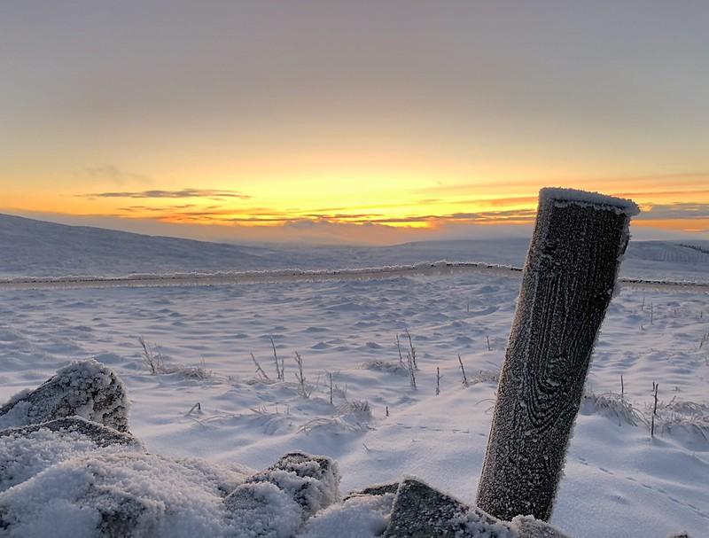 Snow-covered landscape with a single frost-covered wooden post and rocks in the foreground, against a backdrop of a sunrise or sunset.