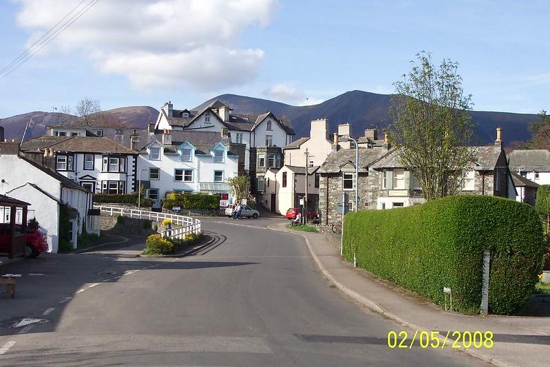 A quaint street with residential buildings in a small town, with green hedges and trees, set against a backdrop of hills and a partly cloudy sky. The date "02/05/2008" is displayed in the corner.