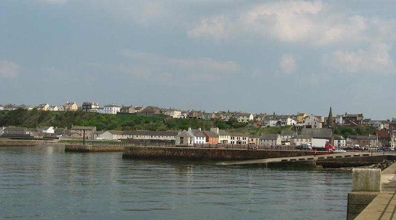 A coastal town with buildings on a hillside, a pier, and a calm body of water in the foreground.