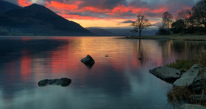 A serene lake at sunset with vibrant red and orange clouds reflected on the water, surrounded by mountains and trees.