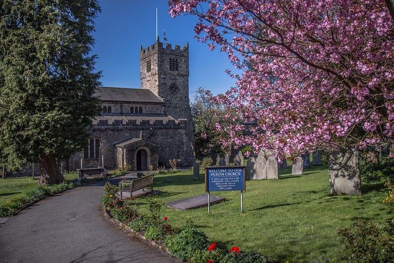 St Andrews Parish Church Sedbergh | Pathway leading to a stone parish church, surrounded by vibrant green grass, benches, tombstones, and a blooming pink tree.