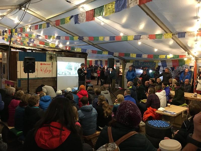 An indoor event with a person speaking on a stage to a seated audience, surrounded by colourful flags and branded signs.