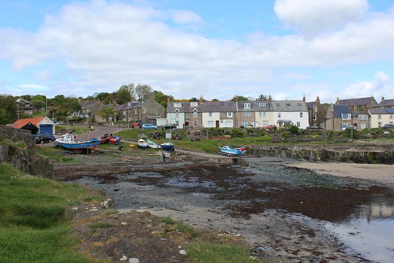 Craster, Northumberland | Small coastal village with a few boats on a grassy area, houses in the background, and a partly cloudy sky.