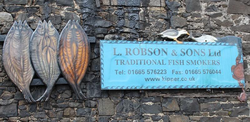 Sign for L. Robson & Sons Ltd Traditional Fish Smokers with a phone number, fax number, and website, mounted on a stone wall with fish decorations.