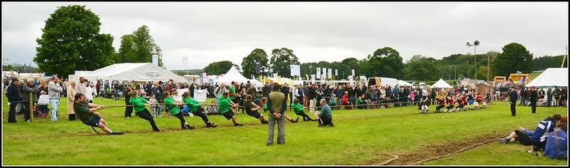 A large crowd watches two teams compete in a tug-of-war on a grassy field with tents and equipment in the background.