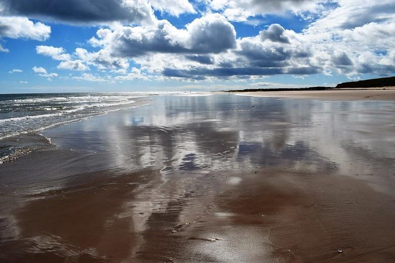 A sandy beach with gentle waves, under a sky filled with clouds and bright blue patches, reflecting on the wet sand.