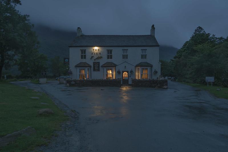 A quaint, illuminated two-story building labeled "The Fish" on a rainy evening, surrounded by trees and a misty backdrop.