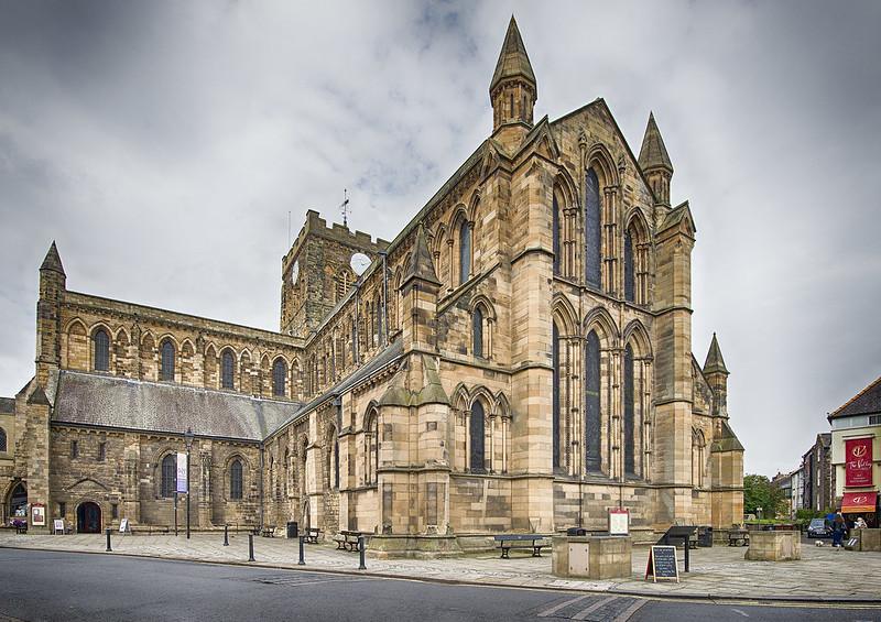 Hexham Abbey, Northumberland | Exterior of a historic stone cathedral with Gothic architectural style, featuring tall arched windows and pointed spires, under a cloudy sky.