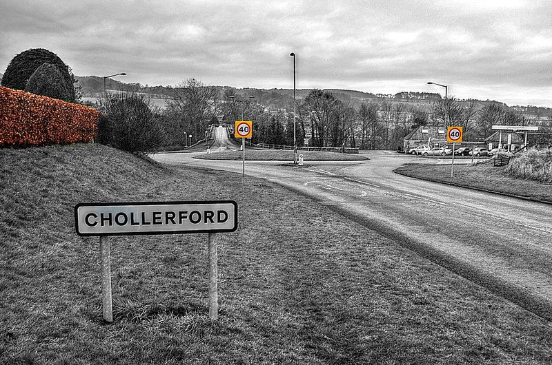 A road sign for Chollerford with a roundabout and 40 mph speed limit signs in the background, surrounded by rural landscape.