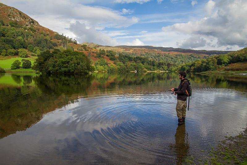 A person standing in a lake fishing, surrounded by lush green hills and a partly cloudy sky.