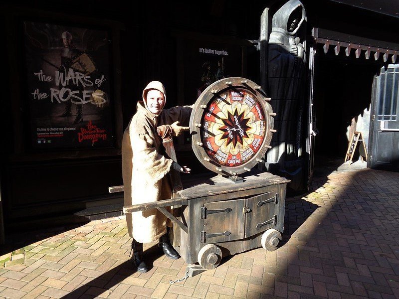 A person in medieval costume stands next to a wheel of fortune on a wooden cart outside, with posters in the background.