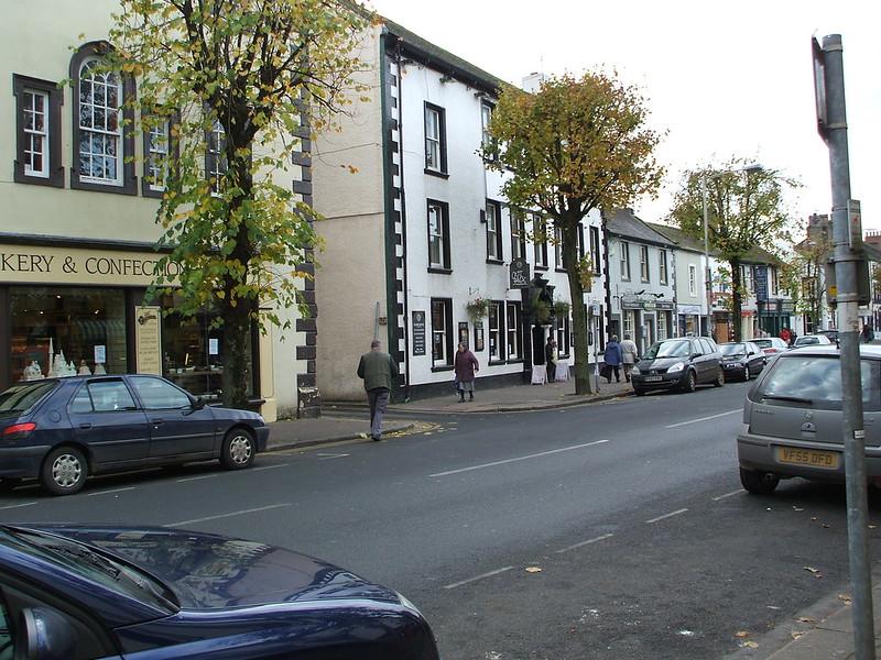 Cockermouth (A594) | Street scene with several people walking on the pavement lined with shops and trees, including a bakery and a confectionery shop.