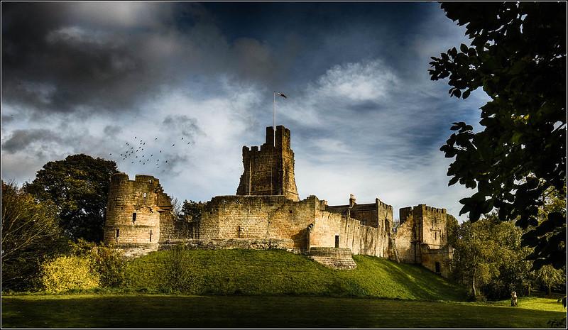 Prudhoe Castle, Northumberland | Medieval castle ruins on a grassy hill under a cloudy sky with some trees in the foreground.