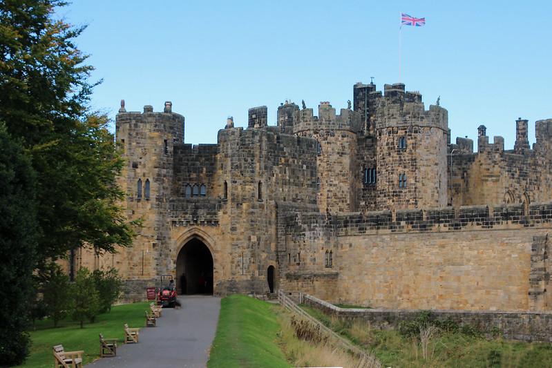 Alnwick Castle with a Union Jack flag flying above, surrounded by greenery and a pathway with benches.