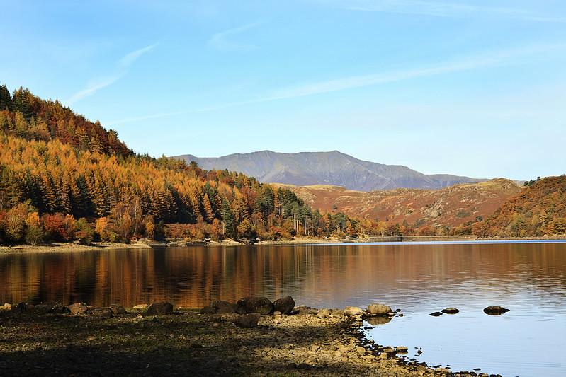 Thirlmere, Cumbria | A tranquil lake surrounded by trees with autumn foliage, and mountains in the background under a clear blue sky.