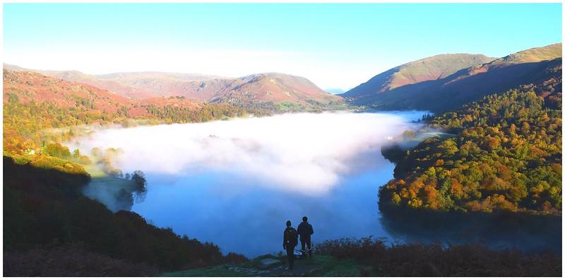 Two people overlook a mist-covered lake surrounded by autumn-coloured hills and mountains.