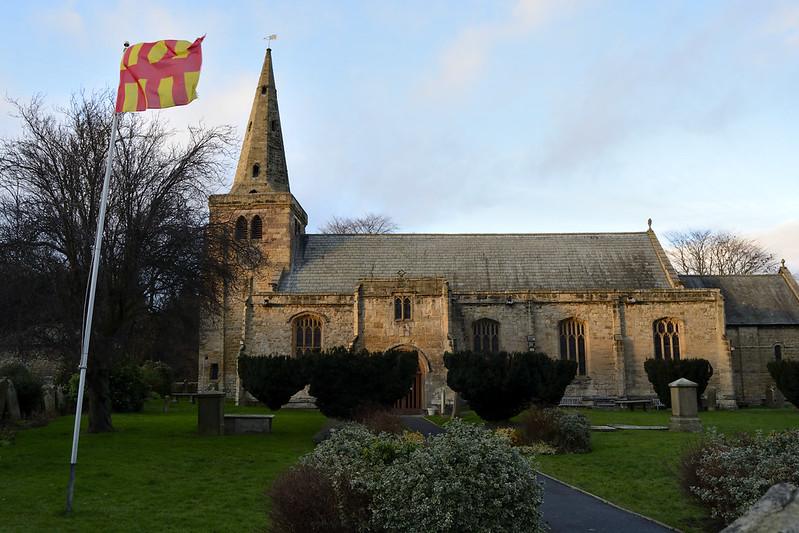 Stone church with a pointed spire, surrounded by a green lawn and trees, with a red and yellow flag on a flagpole in the foreground.