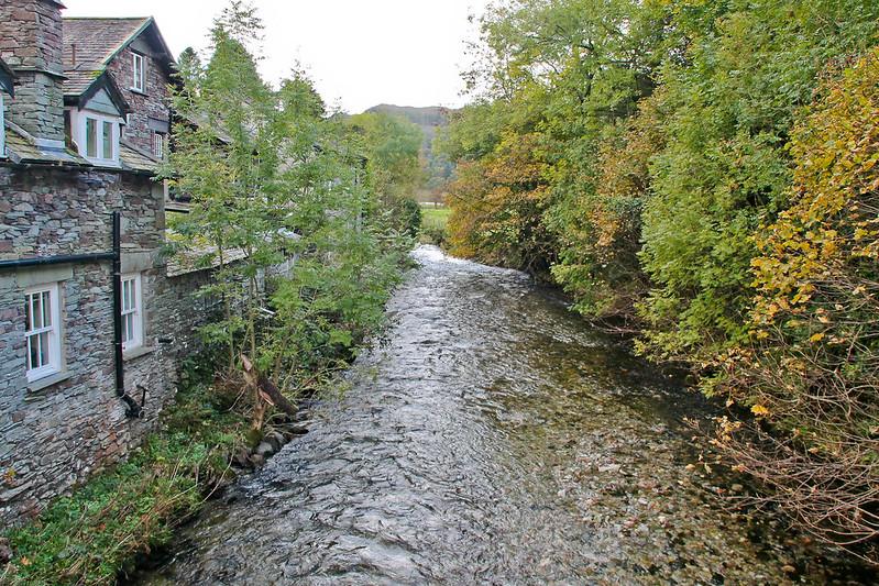 Grasmere, A small river flowing beside a stone house with tall trees lining the riverbank on a cloudy day.