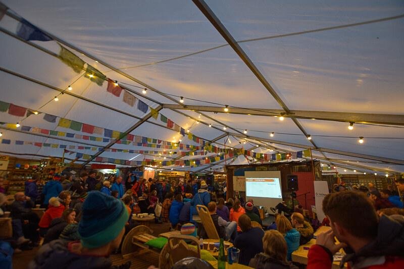 Kendal Mountain Festival, Cumbria | A large group of people gathering inside a tent with hanging colourful flags and string lights, seated at tables and on the ground, facing a screen showing a presentation.