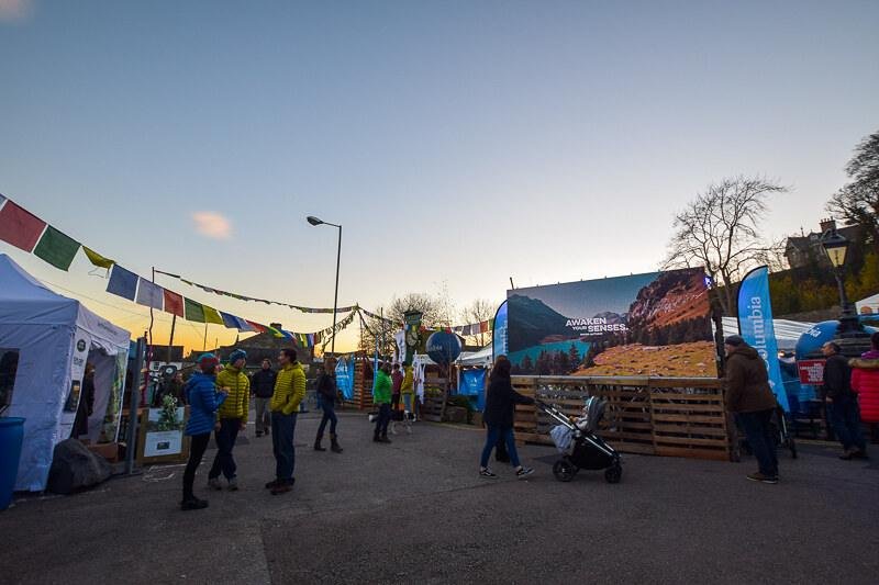 Outdoor festival with booths, people walking, and colourful flags hanging overhead during sunset.