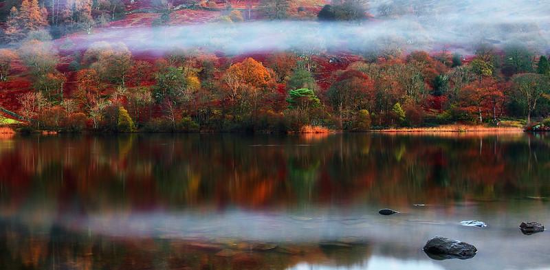 A misty lake with autumnal trees and vibrant foliage reflecting on the calm water, with some rocks in the foreground.