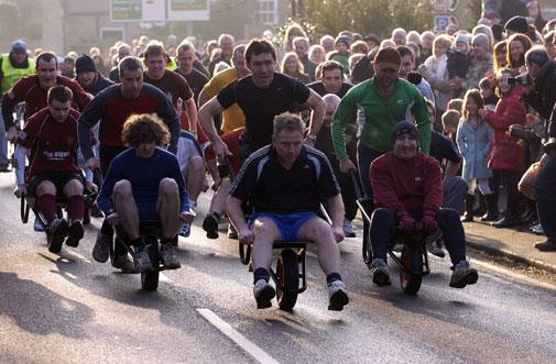 Ponteland Wheelbarrow Race 2009 | A group of men participating in a wheelbarrow race on a street lined with spectators.