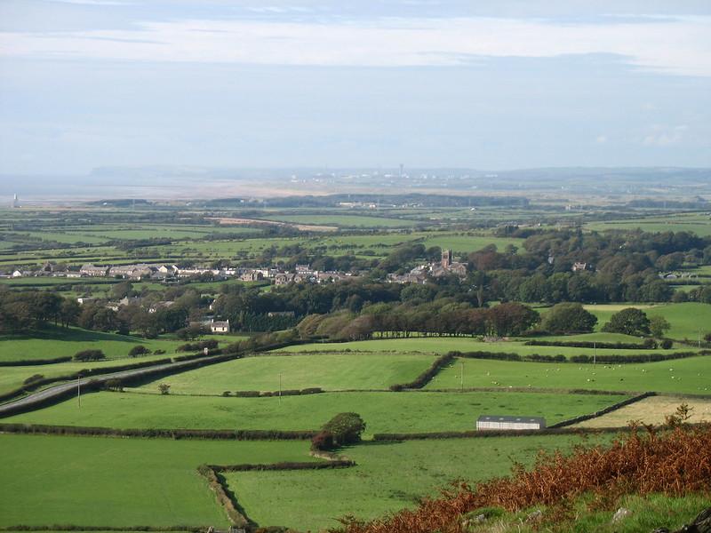 Aerial view of a lush green countryside with variously partitioned fields, a small village with houses and trees scattered throughout, and a distant shoreline with industrial structures visible on the horizon.