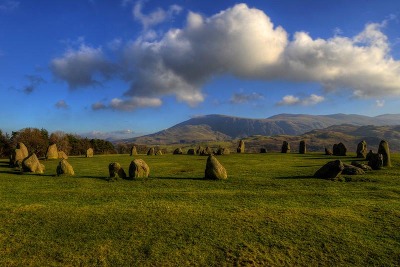 CASTLERIGG STONE CIRCLES | Ancient stone circle on a grassy field with mountains in the background under a partly cloudy sky.