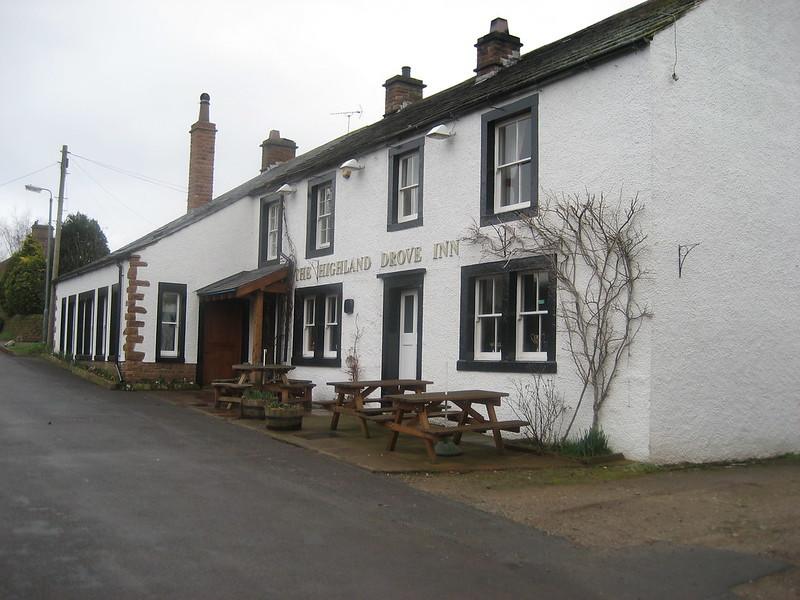 A white-painted building with black window frames and wooden picnic tables in front, identified as "The Highland Drove Inn."