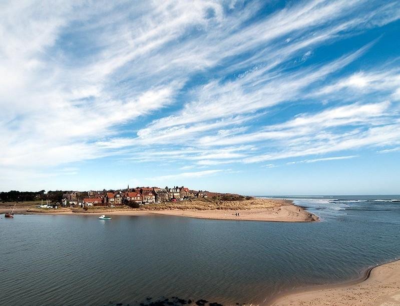 A small coastal village with red-roofed houses overlooking a sandy beach and calm inlet under a blue sky with scattered clouds.