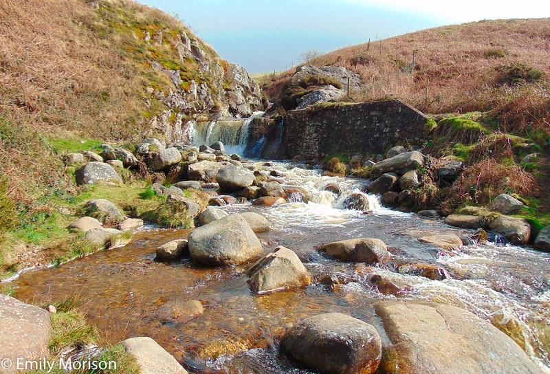 Bootle, Cumbria | A small waterfall cascading over rocks into a stream, surrounded by hilly terrain adorned with grasses and shrubs.