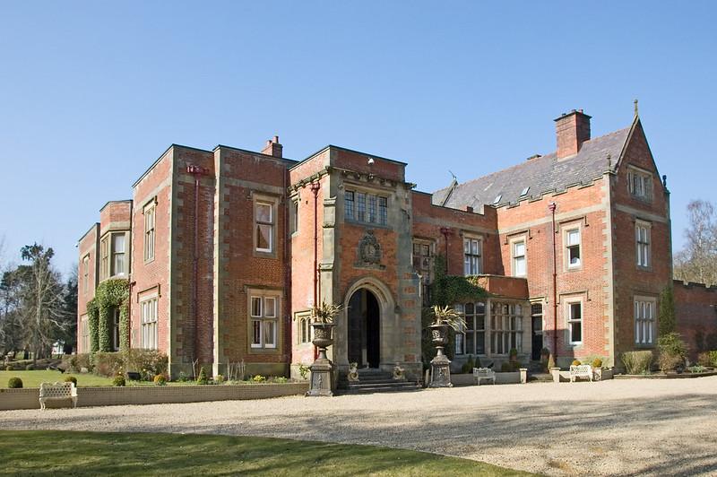 Otterburn Hall | Large, red-brick manor house with ornate stone detailing, large windows, and a grand arched entrance, set against a clear blue sky.