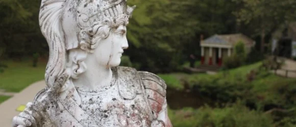 A weathered stone statue of a helmeted figure in armour, set in an outdoor garden with a small building in the background.