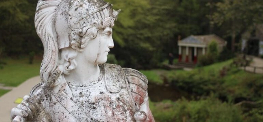 A weathered stone statue of a helmeted figure in armour, set in an outdoor garden with a small building in the background.