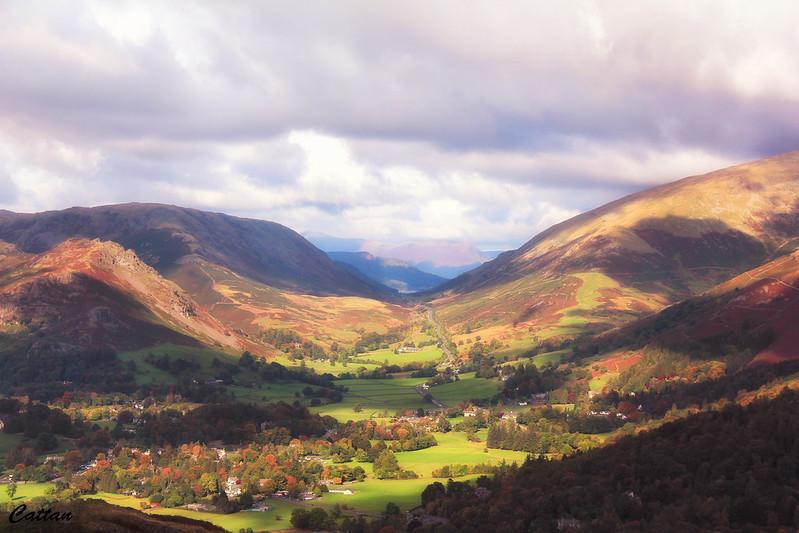 A picturesque valley with scattered houses, lush green fields, and distant mountains under a partly cloudy sky.