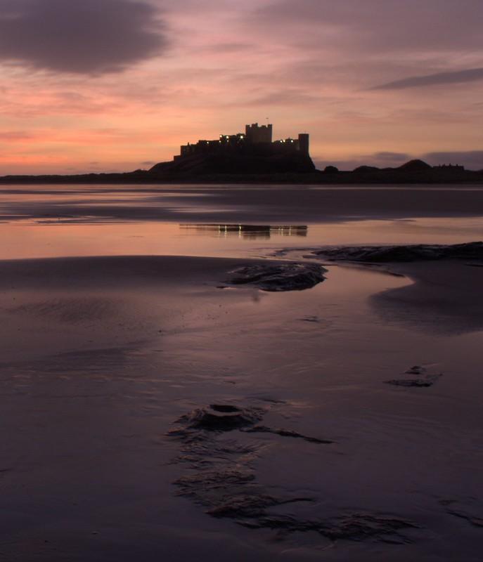 Silhouette of a castle on a hill at sunset, reflecting in wet sand on a beach.