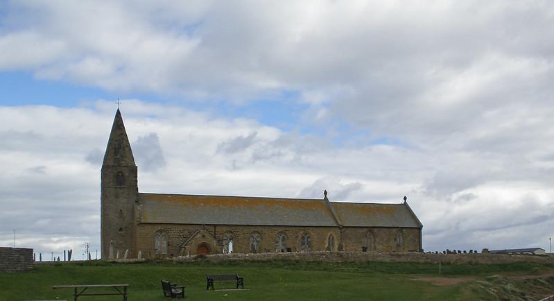 Stone church with a tall, pointed steeple set against a cloudy sky, surrounded by grassy land with benches in the foreground.