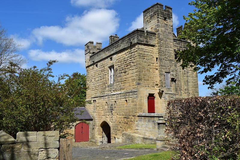 Stone building with red doors and arched entrance surrounded by trees and bushes under a blue sky.