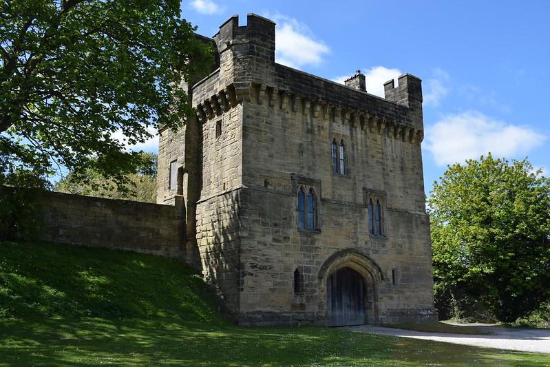 Morpeth Castle, Northumberland | Stone gatehouse with crenellations, arched doorway, and medieval-style windows, surrounded by greenery and under a blue sky with some clouds.