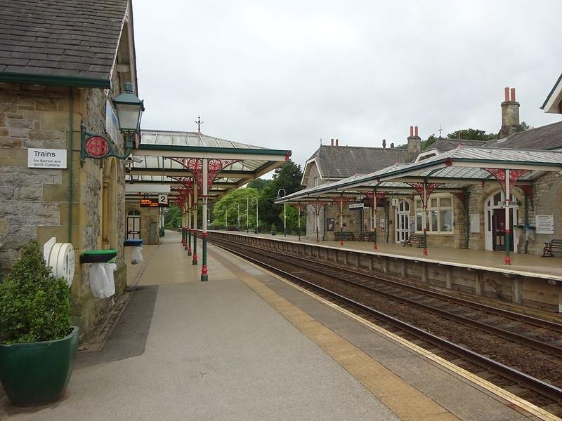 An empty train platform with a Victorian-style railway station, featuring covered walkways, stone buildings, and a sign indicating trains to Barrow and North Cumbria.