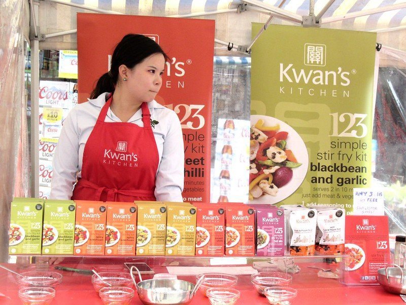 A woman wearing a red Kwan's Kitchen apron stands at a booth displaying various packaged stir fry kits.