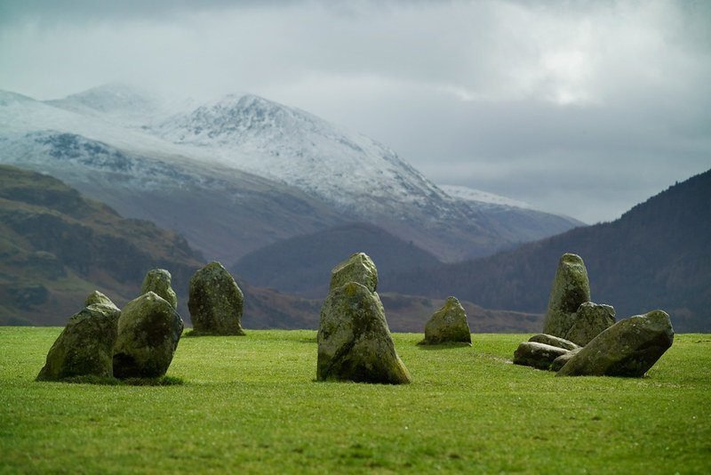 Stone circle on a grassy field with snow-capped mountains in the background.