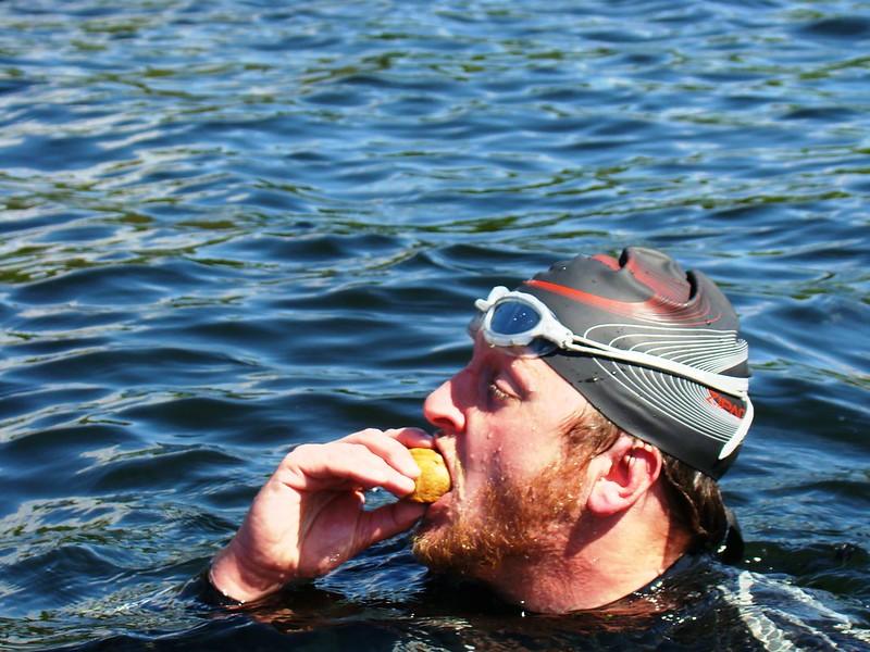 A swimmer wearing a black swim cap and goggles eating a snack while in the water.