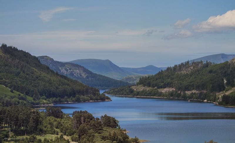 A tranquil lake surrounded by forested hills and mountains under a partly cloudy sky.