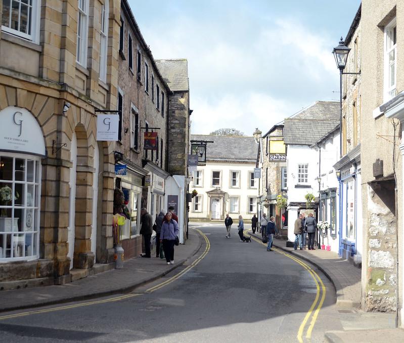 Kirkby Lonsdale, A Narrow street with people walking, bordered by historic stone buildings, signs for shops, and inns, with a larger white building in the background under a cloudy sky.