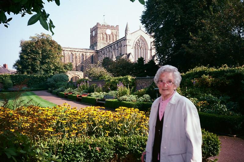 An elderly woman standing in a garden with a large gothic-style church in the background.