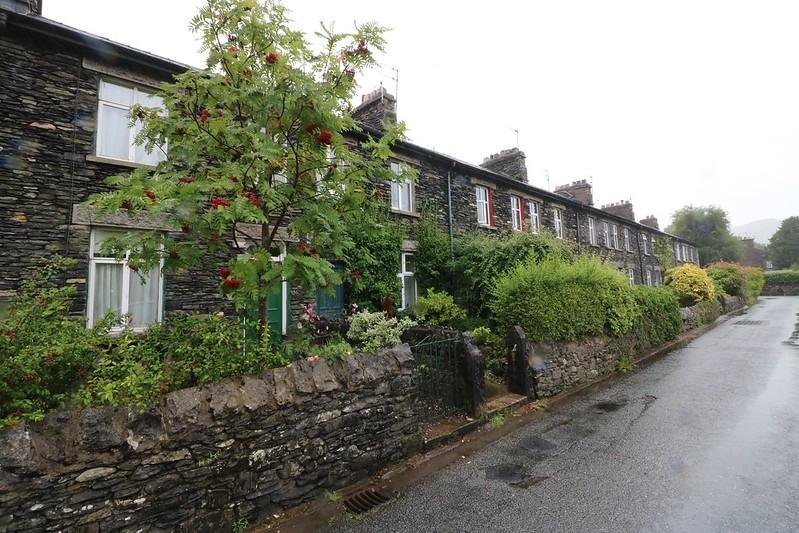 Row of traditional stone terraced houses on a rainy day, with greenery and a tree bearing red berries along the front gardens, beside a wet tarmac road.